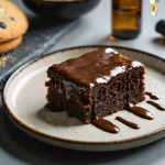 Close-up 45-degree view of a fudgy brownie with ganache on a ceramic plate beside an amber dropper bottle, with cooling cookies, a mixing bowl, and a blurred vape pen in a home kitchen background.
