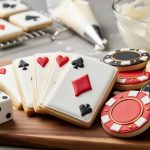 Close-up of casino-themed sugar cookies on a wooden board—white playing-card cookies with red and black suit symbols, round poker chip cookies with subtle gold accents, and dice cookies—lit by soft window light with a blurred cooling rack, piping bag, and icing bowl in the background.