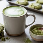 Ceremonial matcha latte in a mug with freshly baked green matcha cookies and a bowl of matcha powder on a countertop.